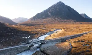 Majestätische Berge der Schottland Highlands, ein Fluss schlängelt sich durch das goldene Gras, überquert von einer Brücke.