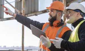 Dwóch mężczyzn w kaskach i kamizelkach odblaskowych omawia projekt na budowie. Jeden wskazuje palcem, drugi trzyma tablet. To przykład dbałości o sluzba bhp.