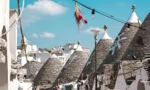 Scorci di Alberobello con i caratteristici trulli, alcuni decorati con simboli bianchi, sotto un cielo azzurro. Una scalinata cuori conduce tra le abitazioni uniche.
