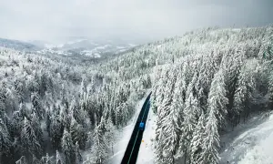Zimowy krajobraz z ośnieżonym lasem i drogą, po której jedzie niebieski autobus.