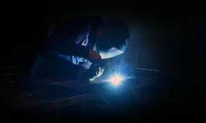 Spawacz w masce ochronnej używa elektrody do żeliwa, tworząc jasny łuk świetlny i dym.