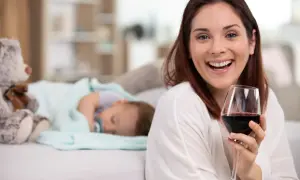 Uśmiechnięta kobieta z kieliszkiem czerwonego wina, podczas gdy dziecko śpi obok. Temat: karmienie piersią a alkohol.