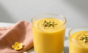 Två glas med krämig, gul indisk yoghurtdryck, toppad med hackade pistagenötter. En rosa duk och små blomformade dekorationer syns i bakgrunden.