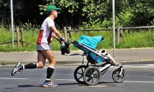 Mężczyzna w zielonej czapce i sportowym stroju bierze udział w biegu z wózkiem. Dziecko w wózku ma na sobie białe skarpetki i sandałki.