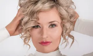 Kobieta z kręconymi blond włosami, jej spojrzenie jest nonszalanckie, a dłonie unoszą się ku głowie.