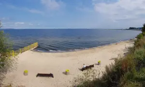 Plaża dla psów z łagodnym zejściem do morza. Na piasku ławki i kosze. W oddali widać falochron.