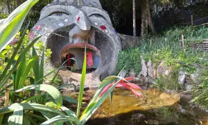 Scultura di una testa gigante con bocca spalancata nel giardino botanico Gardone Riviera, con piante rigogliose e uno stagno.