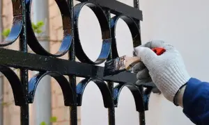 Ręka w rękawicy maluje pędzlem czarne, ozdobne przęsła ogrodzeniowe, odnawiając ich wygląd.