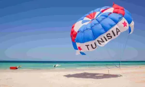 Kolorowy spadochron z napisem "TUNISIA" unosi się nad plażą, zapowiadając wspaniałe dżerba loty. W tle turkusowe morze i piaszczysta plaża.