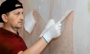 Mężczyzna w czapce i rękawiczkach zrywa starą tapetę, odsłaniając wzorzystą powierzchnię.