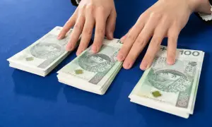 Dłonie nad stosem banknotów 100 zł. Symbolizuje to darowiznę między małżonkami, wsparcie finansowe lub oszczędności.