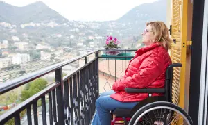 Donna in carrozzina su un balcone, con vista su un paesaggio collinare. Un'immagine che evoca la necessità di un piano nazionale non autosufficienza.