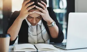 Kvinna med händerna i håret, blicken sänkt över en öppen bok. Känslan av stress på jobbet är påtaglig. En kaffekopp och laptop finns i närheten.