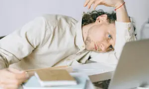 Giovane uomo con astenia neurologica, appoggiato alla scrivania, sguardo perso nel vuoto, laptop aperto.