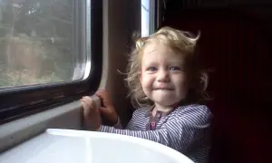 Uśmiechnięta dziewczynka z blond włosami cieszy się wycieczką pociągiem, patrząc przez okno na mijany las.