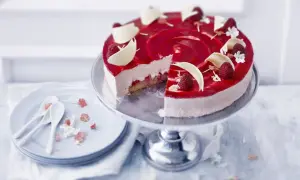 En vacker hallon cheesecake med hallonspegel, vita chokladdekorationer och färska hallon. Serveras på ett tårtfat med en skiva borttagen.