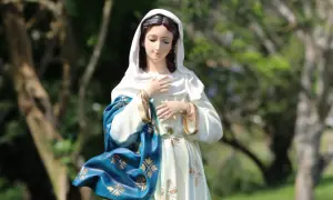 Figurka Matki Boskiej w błękitnym płaszczu ze złotymi zdobieniami. W tle drzewa. Idealna na 3 maja święto kościelne.