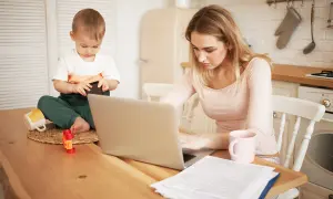Młoda mama pracuje na laptopie, podczas gdy jej małe dziecko bawi się obok. Czy na urlopie macierzyńskim można pracować? Tak, często jest to możliwe.