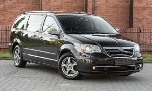 Czarny Lancia Voyager stoi na brukowanym podjeździe przed ceglanym budynkiem.