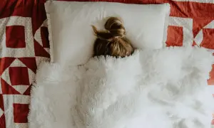En person med en tofs i håret vilar på en kudde under ett fluffigt täcke. Kanske känner sig personen lite hängig, som vid feber gravid.