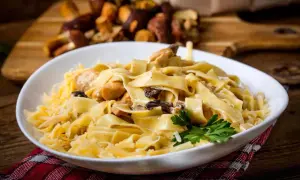 En skål med krämig pasta med kyckling och svamp, toppad med riven ost och en persiljekvist. I bakgrunden syns färska karljohanssvampar. En härlig pasta porcini-rätt.