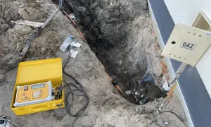 Wykonanie przyłącza gazu od skrzynki do domu. Koparka odsłoniła rurę gazową, obok leży żółta walizka z aparaturą pomiarową.