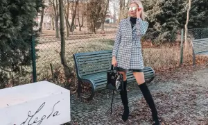 Stylowa kobieta w wełnianym płaszczu w pepitkę, czarnych kozakach i okularach przeciwsłonecznych pozuje w parku.