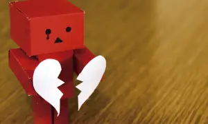 Un robot rojo de origami, con cara triste y una lágrima, sostiene las dos mitades de un corazón blanco. Parece estar a punto de morir de amor.