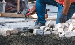 Brukarz w rękawicy i jeansach precyzyjnie układa kamienie, używając młotka. Praca brukarza wymaga cierpliwości i dokładności.