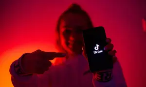 Dłoń wskazuje na ekran telefonu z logo TikToka. W tle czerwone, neonowe światła, tworzące atmosferę jak z klubu z tantiemami.