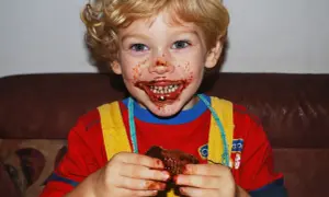 Bambino con cioccolato in bocca e sulle mani, che tiene un pezzo di torta.