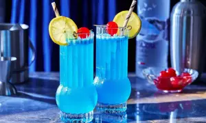 Dwa orzeźwiające drinki Błękitna Laguna, ozdobione cytryną i wiśnią, stoją na barze obok butelki wódki i shakera.