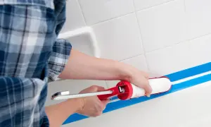 Osoba w kratę zakłada silikon sanitarny w wannie, używając pistoletu i taśmy malarskiej.