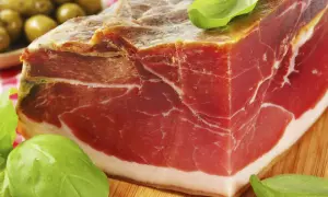 Ein Stück Parmaschinken mit Basilikum und Oliven auf einem Holzbrett.