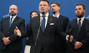 Mężczyzna przemawia przy mikrofonie, otoczony innymi politykami. W tle niebieska ściana z napisem. Konfederacja postulaty.