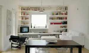 Soggiorno moderno con ampie librerie cartongesso a parete, piene di libri, che incorniciano una finestra. Divano bianco e poltrona nera completano l'arredo.