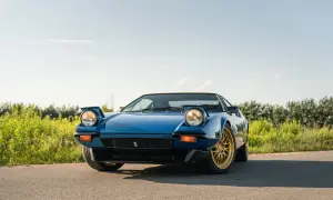 Niebieski De Tomaso Pantera z wysuwanymi światłami i złotymi felgami na tle zieleni i błękitnego nieba.