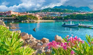 Vue d'une baie turquoise en Sardaigne, avec des maisons roses, un pont en bois et des fleurs roses. Idéal pour savoir que faire en Sardaigne.
