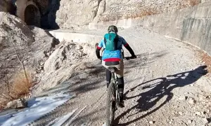 Ciclista in mountain bike su sentiero sterrato, con neve residua e un tunnel scavato nella roccia. Un'avventura tra le **Tignale cascate**.