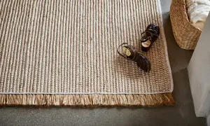 En ljus, flätad matta med fransar ligger på ett betonggolv. Ett par bruna sandaler vilar på mattan.