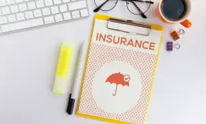 Planowanie ubezpieczenia zdrowotnego bez pracy. Na biurku klawiatura, okulary, kawa i dokument "INSURANCE" z parasolką.