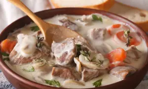 Un plat réconfortant de véritable recette blanquette de veau, avec des morceaux de viande tendre, des champignons et des carottes dans une sauce crémeuse, servi dans un bol en bois avec une cuillère.