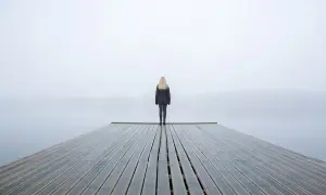 Una persona fredda, con la psicologia che la isola, sta in piedi su un molo di legno in una giornata nebbiosa.