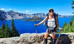 Uśmiechnięta turystka z plecakiem i kijkami, podziwiająca błękitne jezioro. To widok z trasy Pacific Crest Trail, pokazujący, ile się idzie przez takie cuda natury.