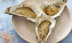 Trois huitres au four, garnies de chapelure dorée, reposent sur un lit de gros sel sur une assiette rose.