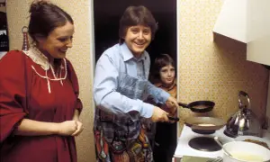 Familie in der Küche, der Vater brät Pfannkuchen nach DDR Rezepten. Ein Kind schaut zu, die Mutter lächelt.