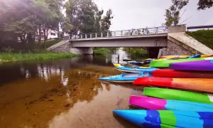 Kolorowe kajaki czekają nad rzeką, idealne na spływ kajakowy Mazury gdzie najlepiej. Most i zieleń tworzą malowniczy krajobraz.