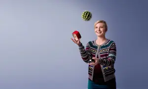 Kvinna jonglerar med en kaktus, en röd väckarklocka och en metallkula. Hon ser glad ut, som om hon klarar allt. Osa 2015.
