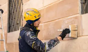 Operaio con casco giallo intento a intonacare muro vecchio con un pennello largo.