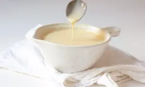 Cuchara vertiendo crema inglesa en un cuenco blanco sobre tela arrugada.
